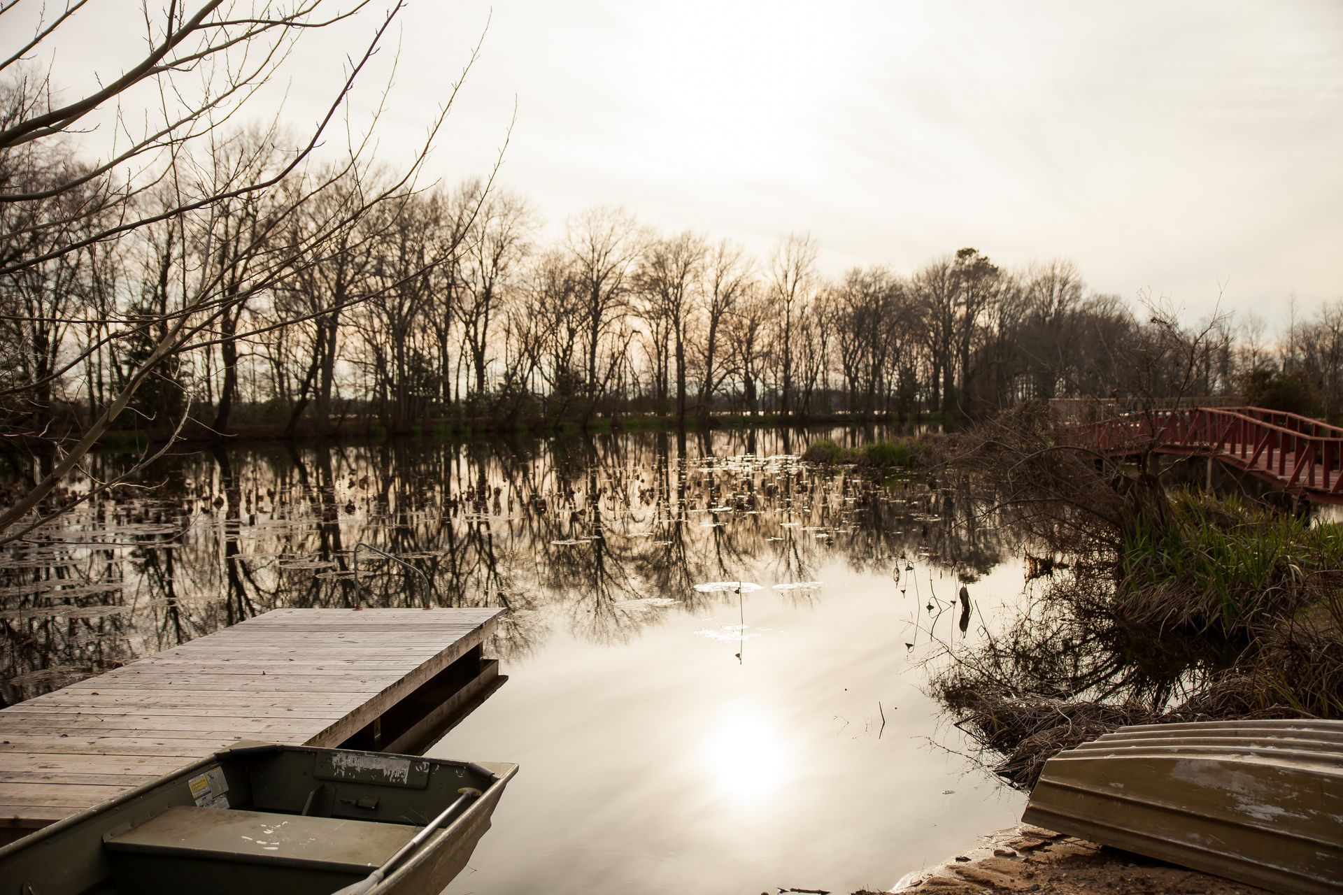 A wooden dock and a small boat sit on a calm lake reflecting trees under a bright, hazy sky at sunset.