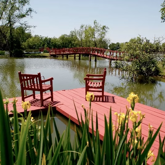 Two red wooden chairs on a matching deck overlooking a pond with a red arched bridge in the background.