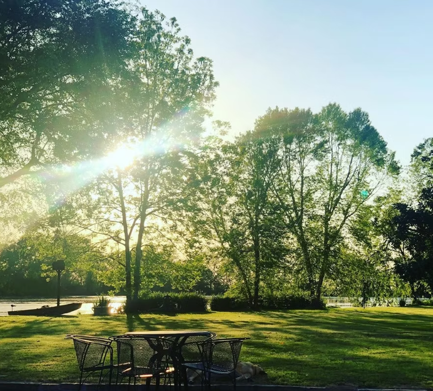 Sunlight filters through trees over a grassy lawn with a metal table and chairs overlooking a river at sunset.