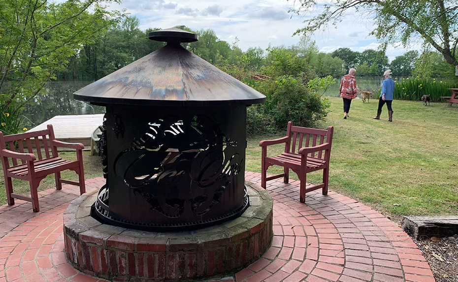 A metal fire pit on a brick patio with two chairs, overlooking a river with two people walking in the background.