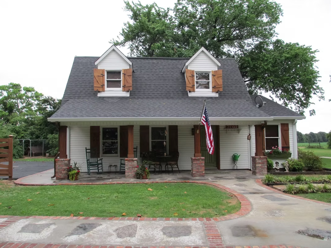 A charming white, one-and-a-half-story house with a front porch, dormer windows, wooden shutters, and a brick walkway.