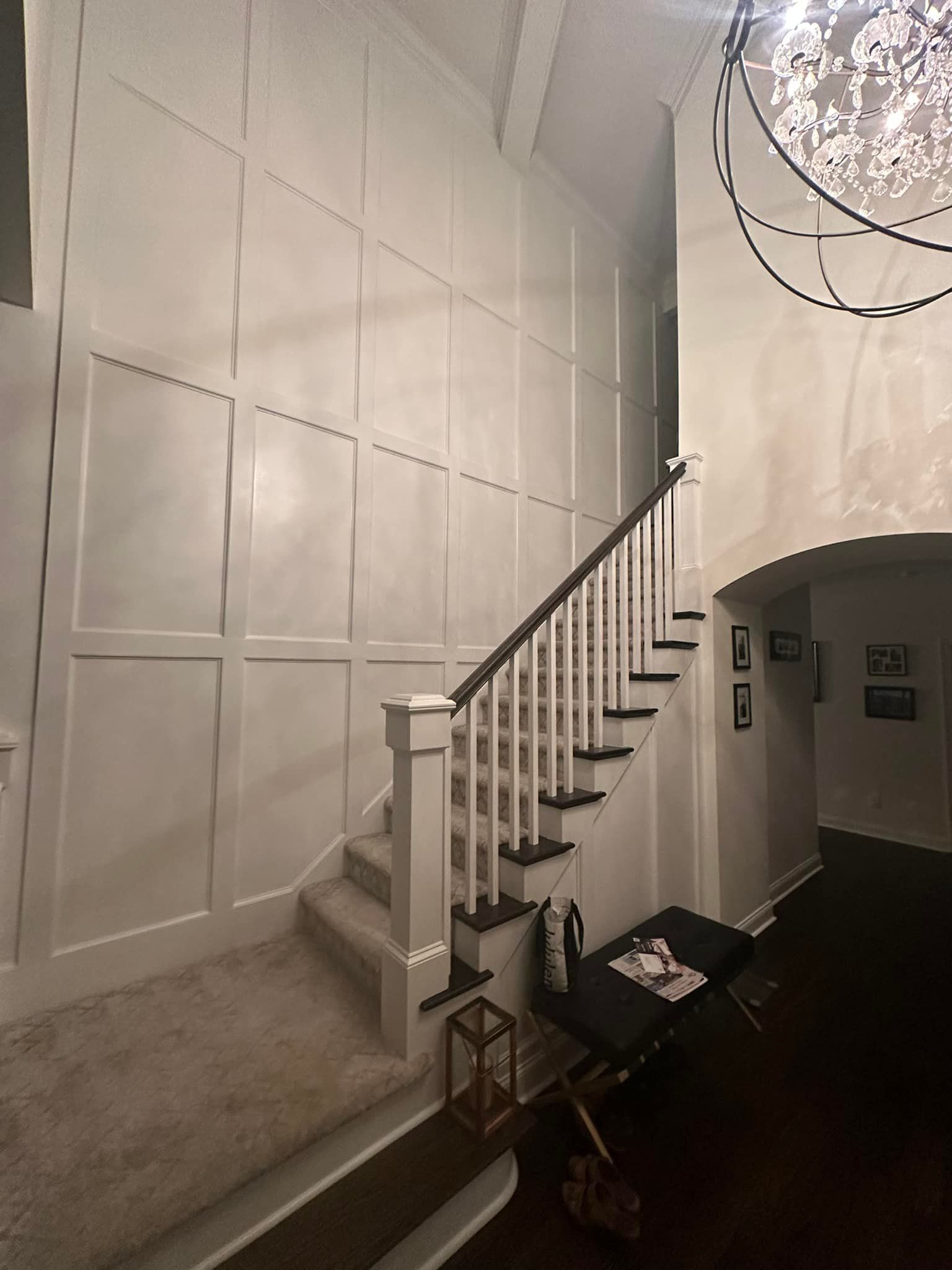 A staircase with white paneling on the wall, leading upwards, carpeted stairs, and a decorative chandelier.