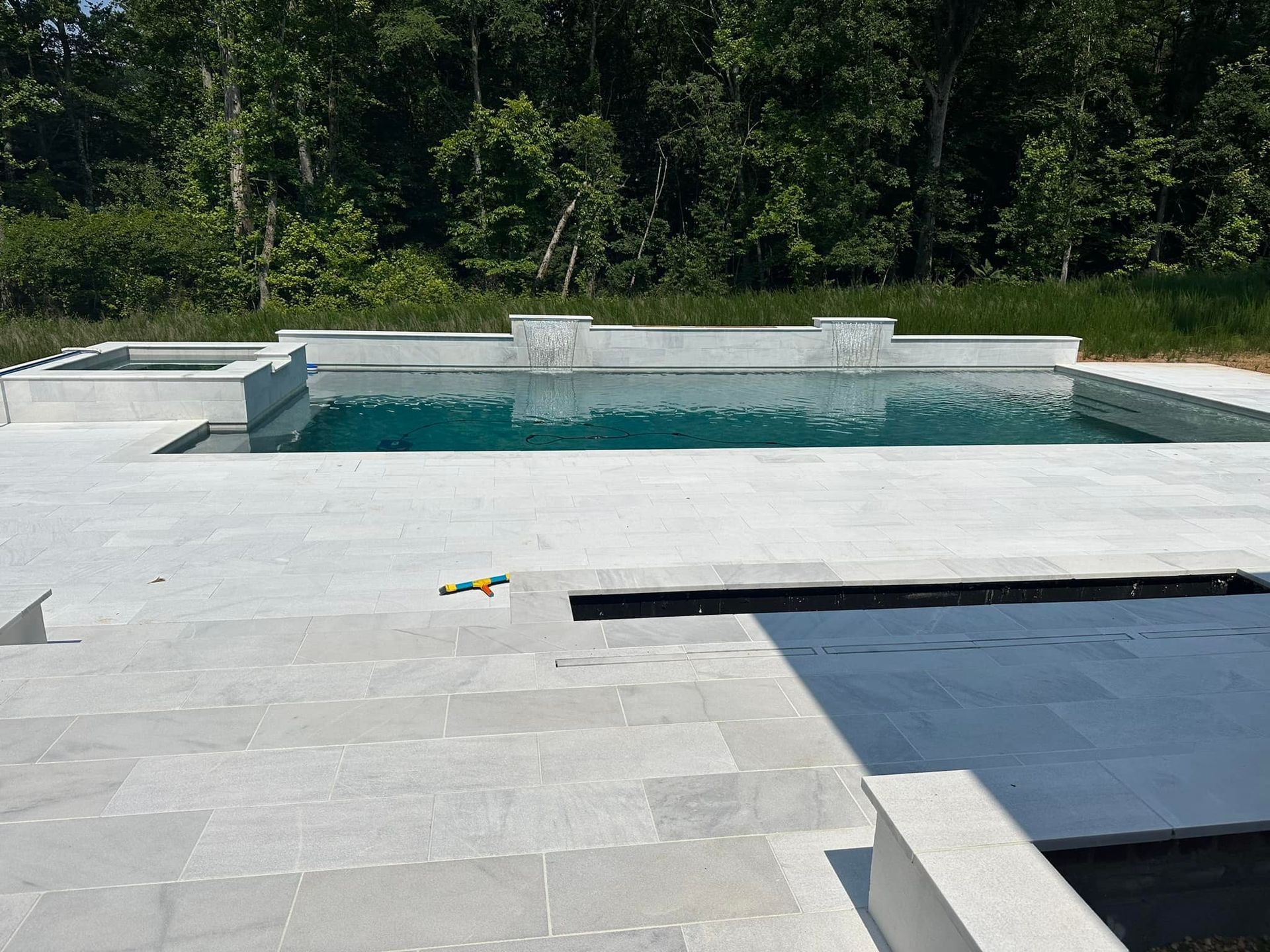 Rectangular pool with gray stone patio, low wall, and trees in the background on a sunny day.