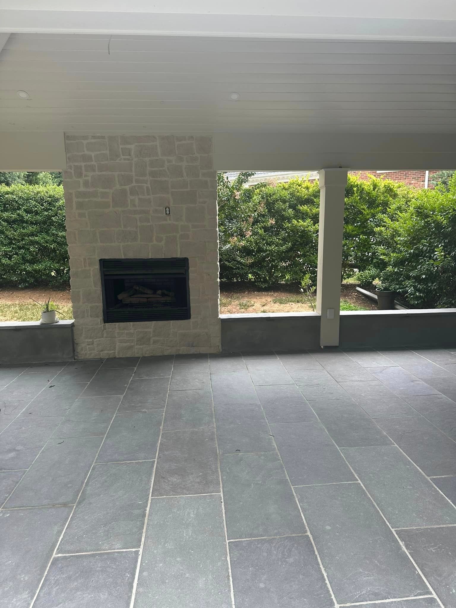 Patio with a stone fireplace, dark tiled floor, and green hedge background.