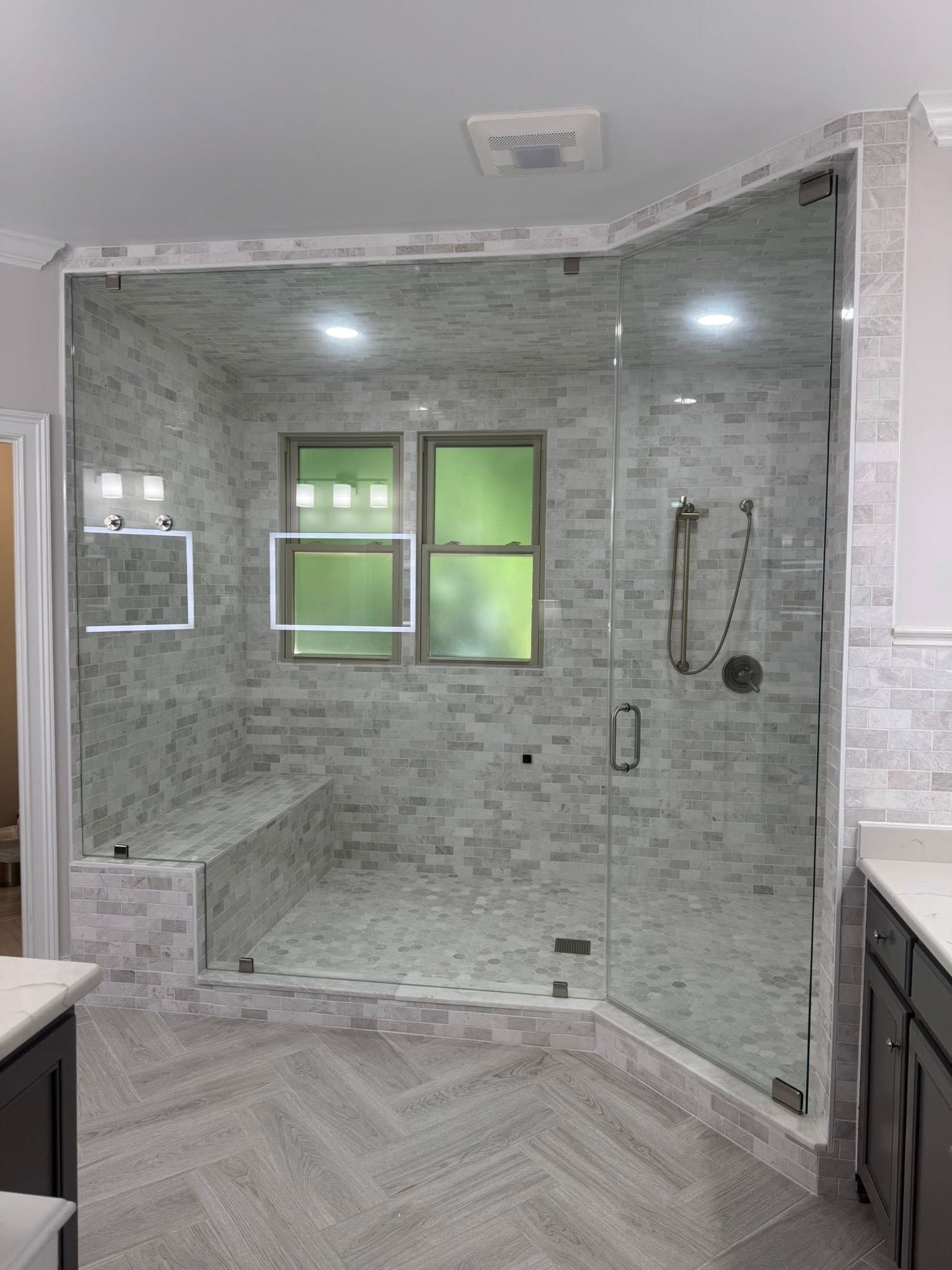 Modern bathroom with glass shower enclosure, gray and white tile, two windows.