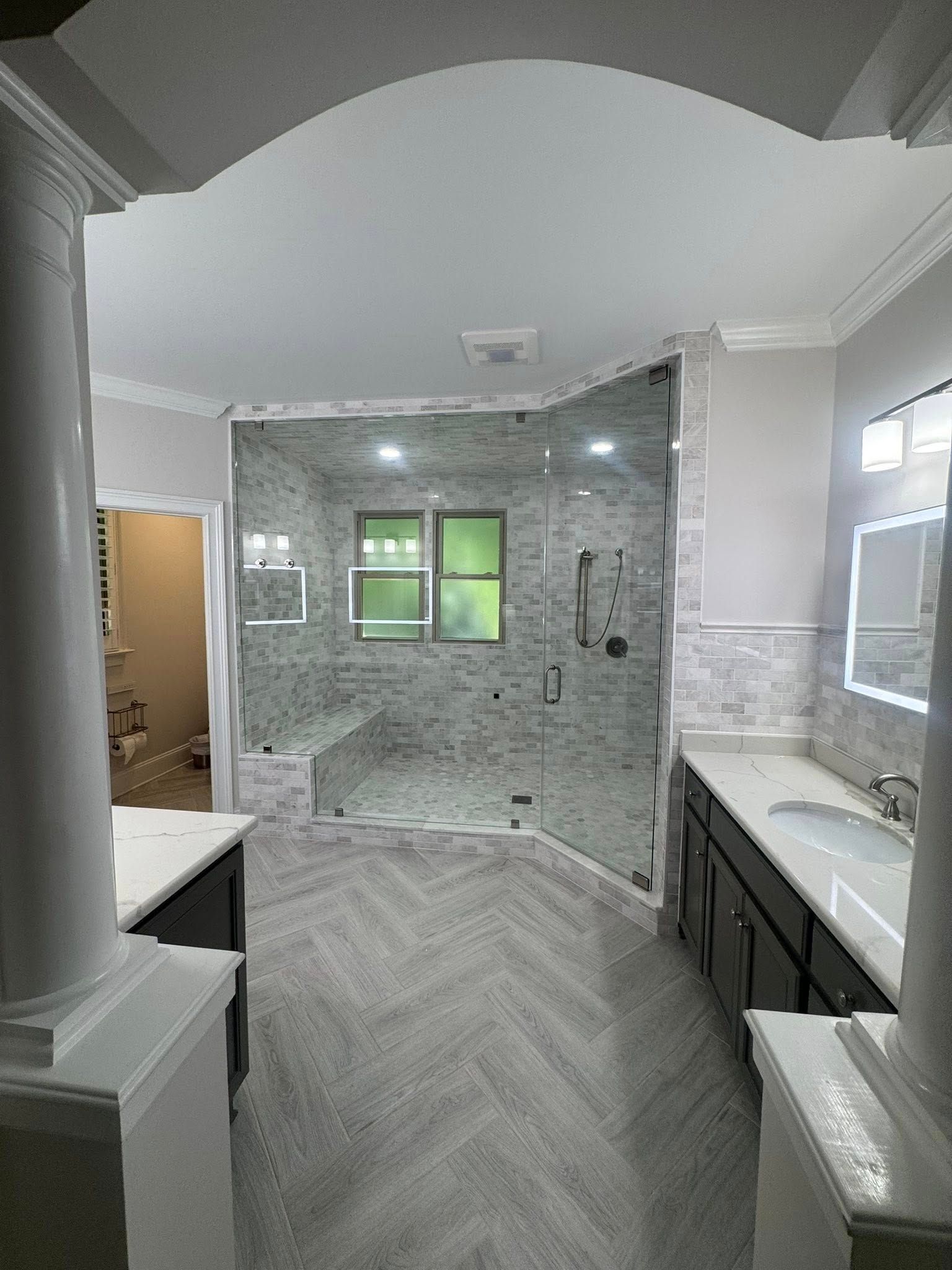 Bathroom with a glass-enclosed shower, two vanities, grey herringbone floor, and archway.