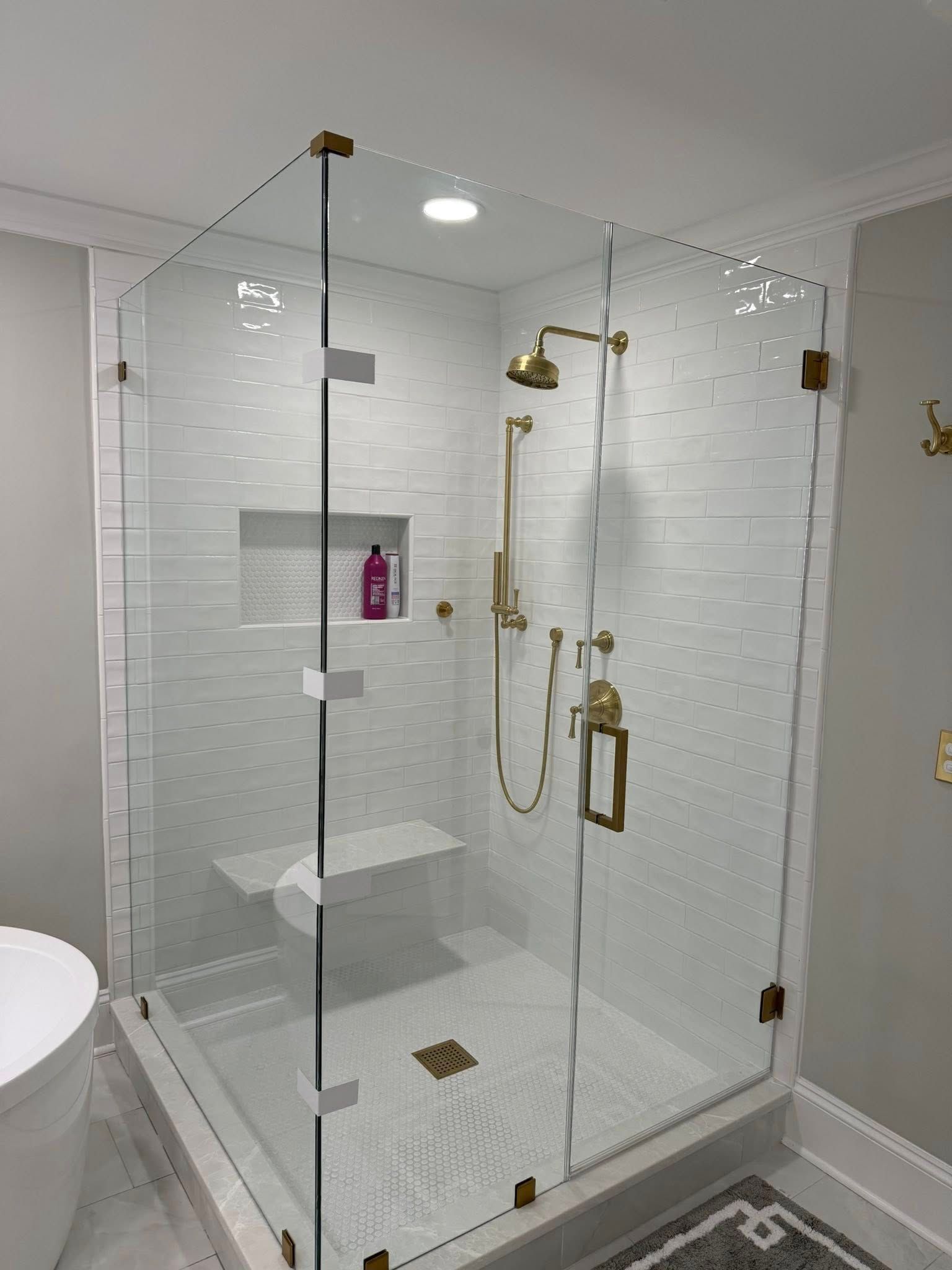 Glass-enclosed shower with gold fixtures and white tile walls. Includes a built-in seat and shampoo niche.