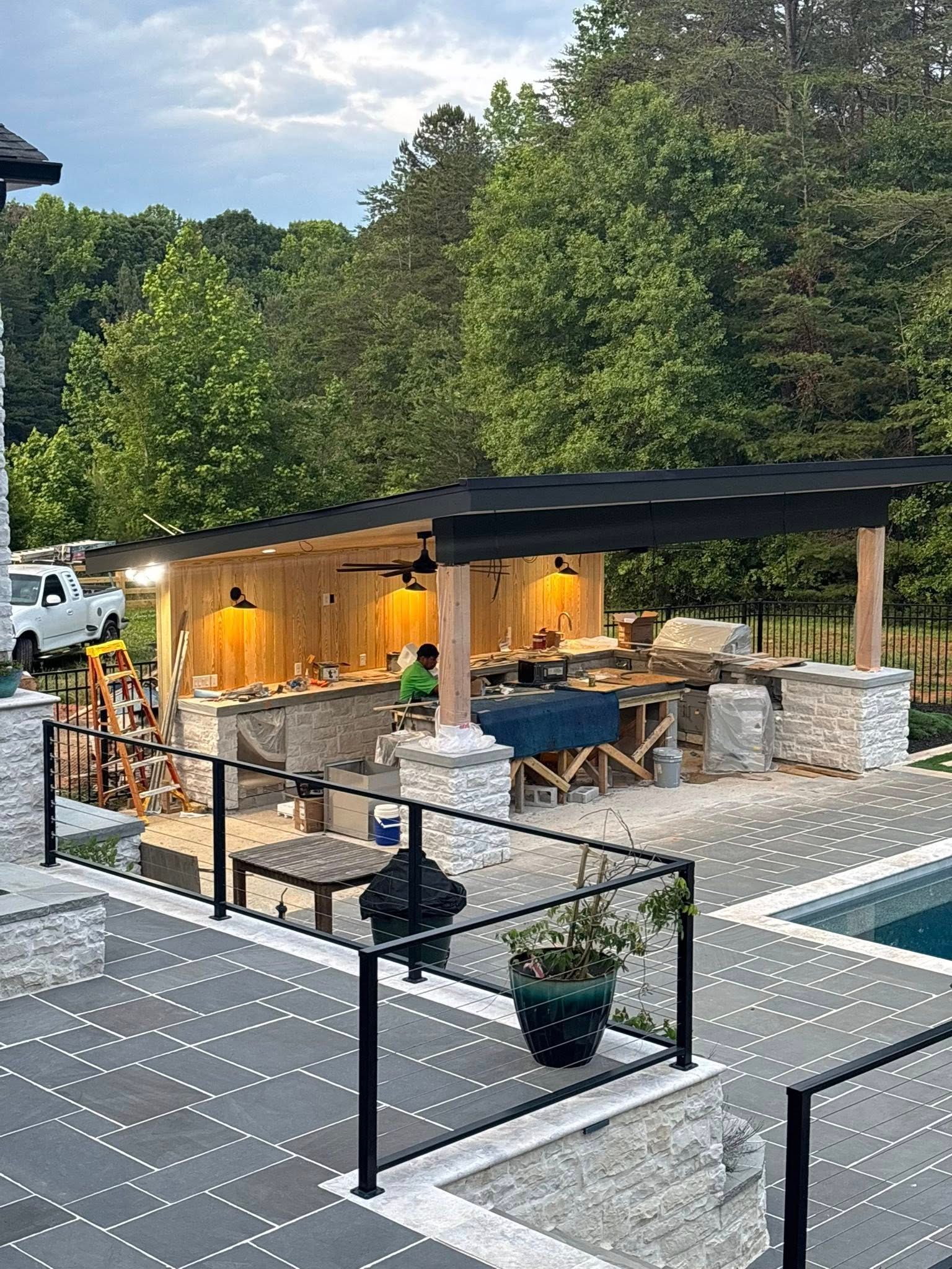 Outdoor kitchen with a stone facade, wooden pergola, and black railing by a pool.