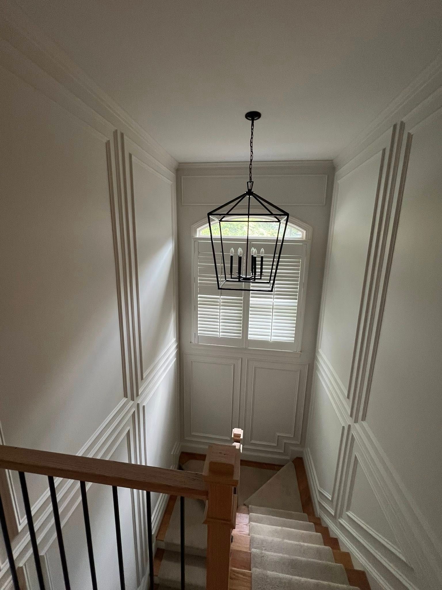 Stairwell with white walls, decorative molding, and a chandelier. Window with shutters at top. Wooden handrail.