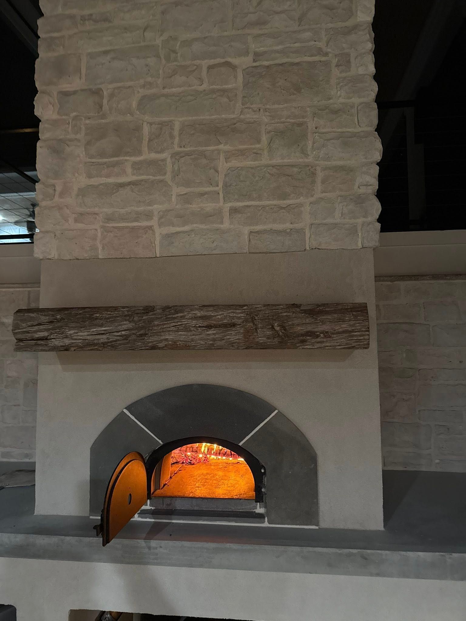 Stone pizza oven with an open door and a wood beam mantel.