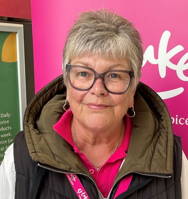 Woman wearing glasses, a pink shirt, and a black vest, smiling. Retail setting, possibly a store.