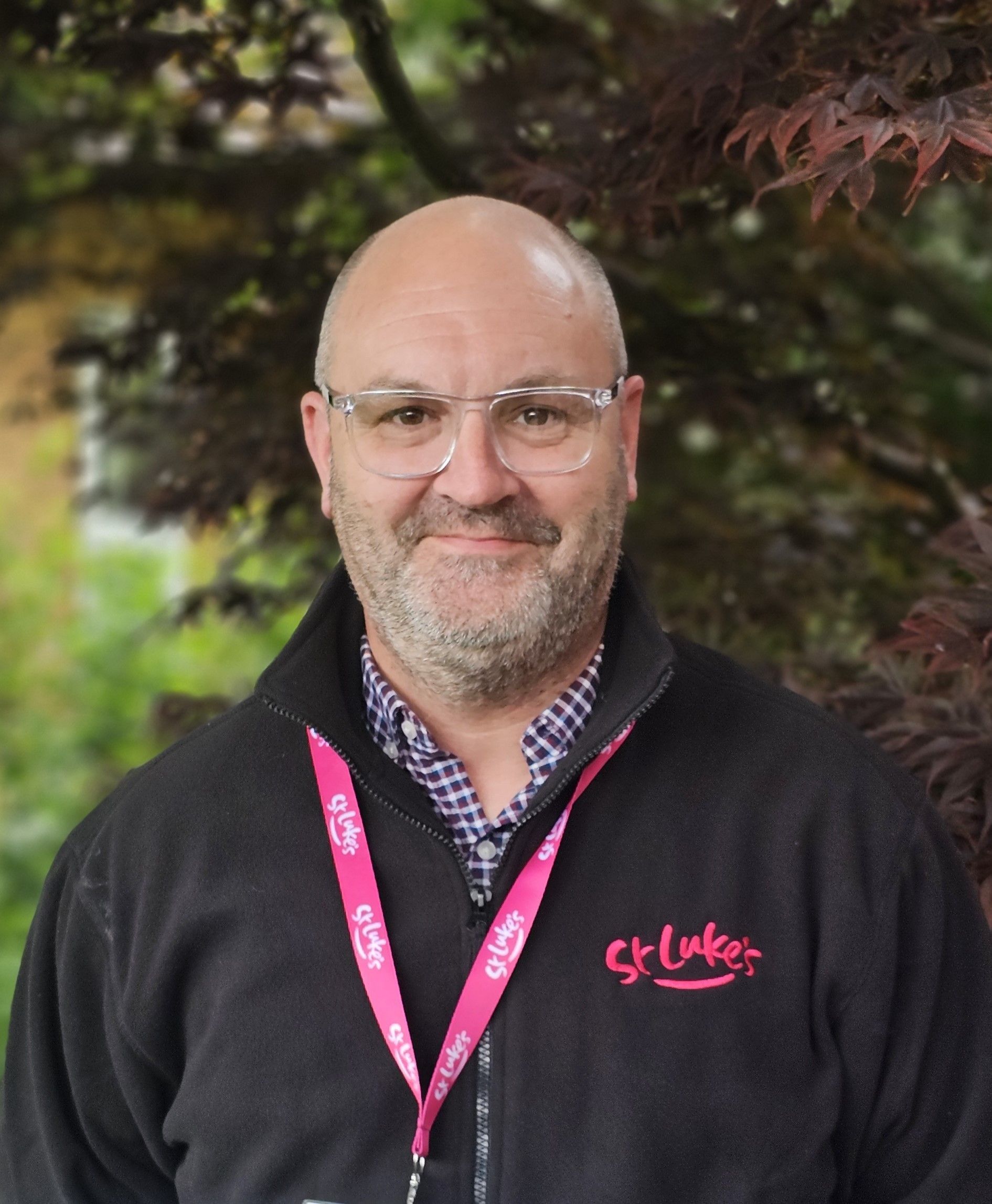 Man with glasses wearing a black jacket and pink lanyard, smiling in front of a tree.