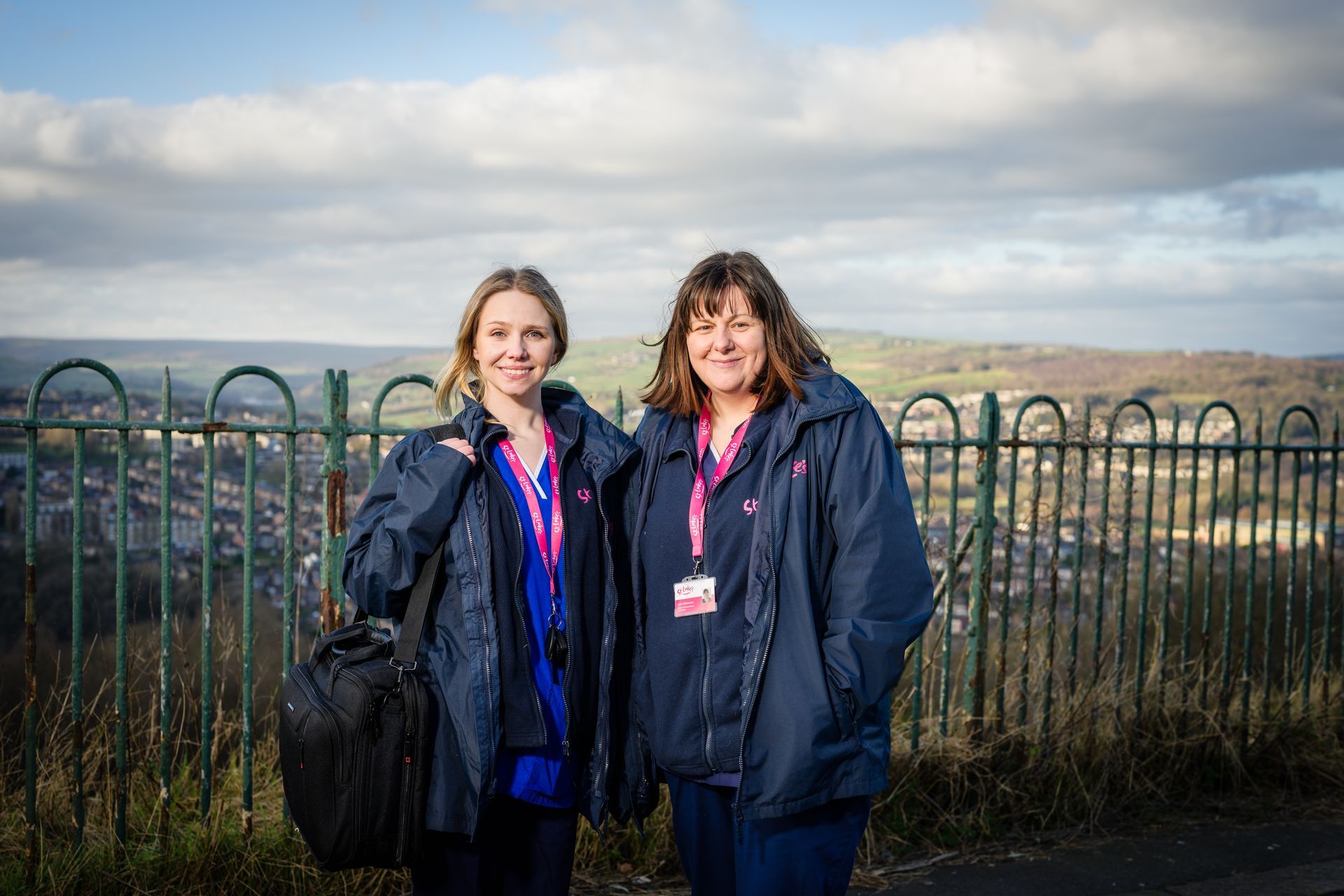A healthcare worker in a blue uniform and mask smiles at a person sitting at a table.