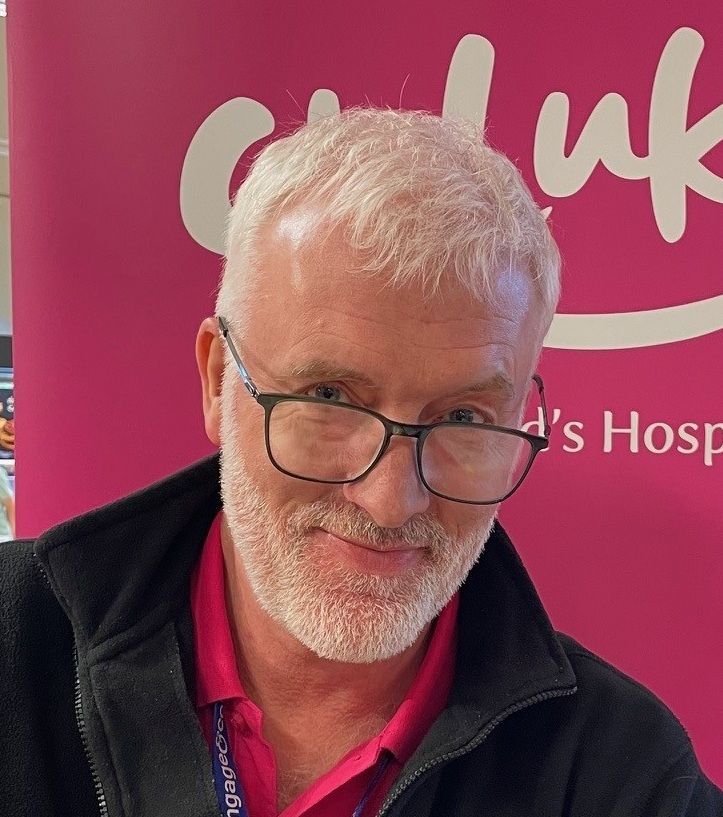 Man with glasses and white hair smiles, standing in front of a pink Child UK Hospital backdrop.
