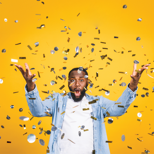 Man with arms raised, mouth open in surprise, surrounded by confetti on yellow backdrop.