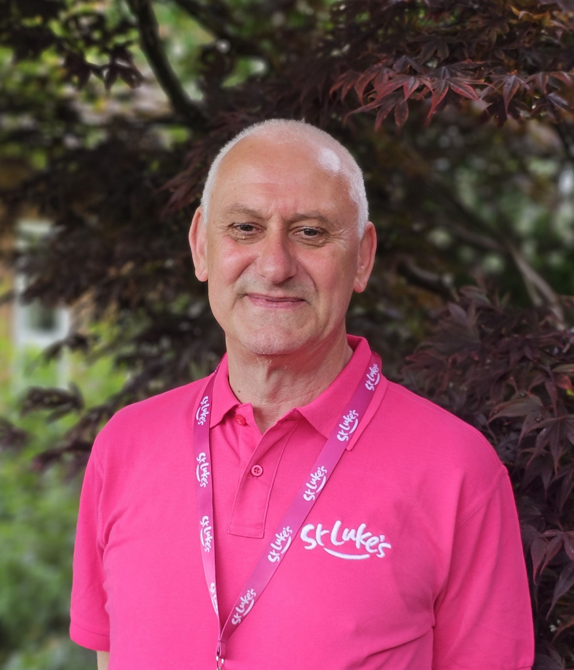 A person wearing a pink polo shirt with a St. Luke’s logo and lanyard, standing in front of dark-leafed foliage.