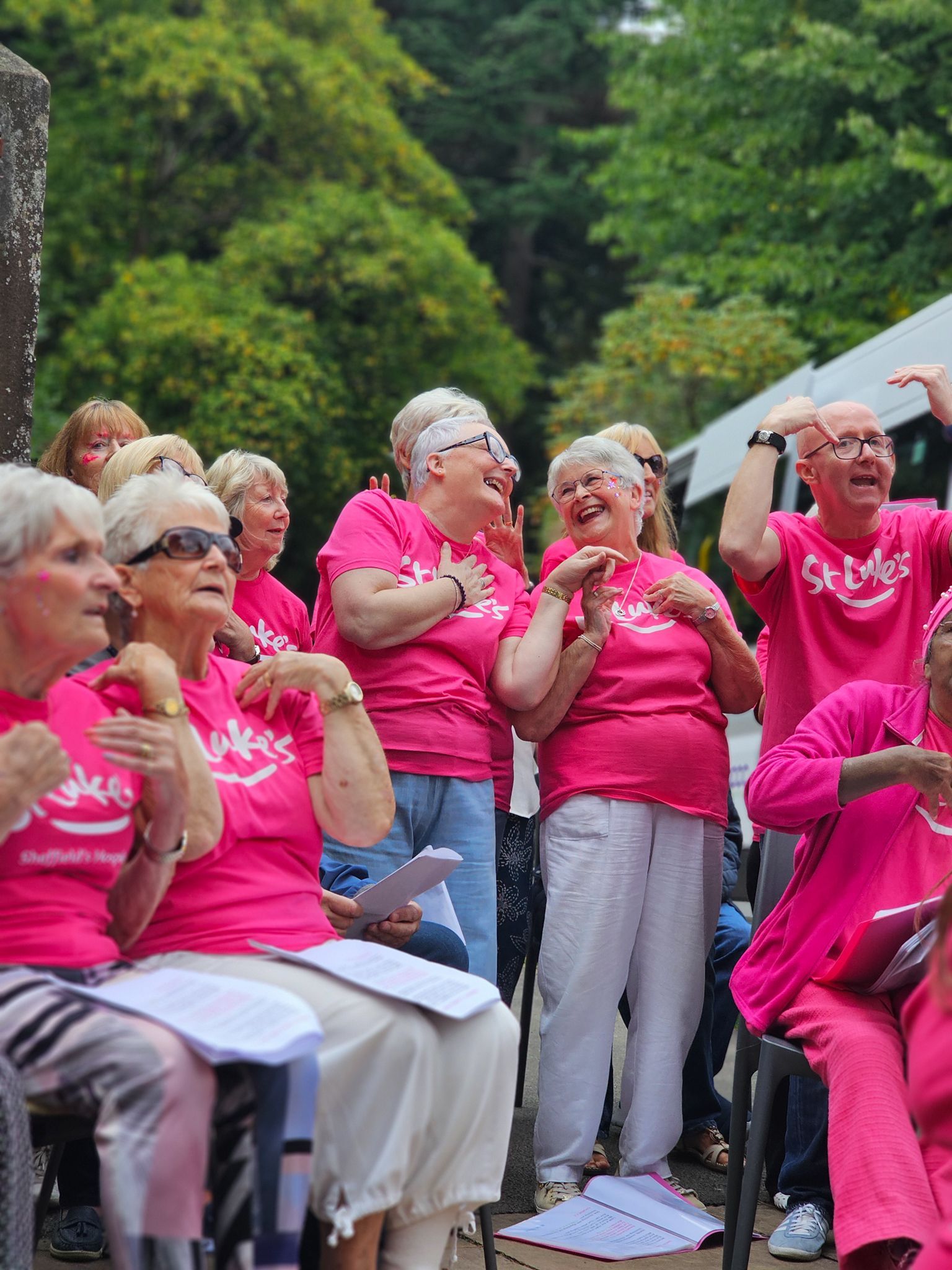 A group of people wearing pink shirts sing outdoors, some laughing and gesturing, amidst green trees.