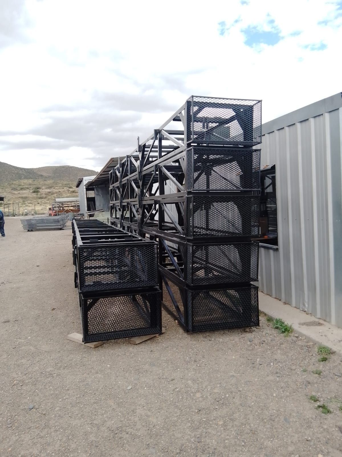 Jaulas de almacenamiento de metal negro apiladas sobre grava cerca de un edificio bajo un cielo nublado.