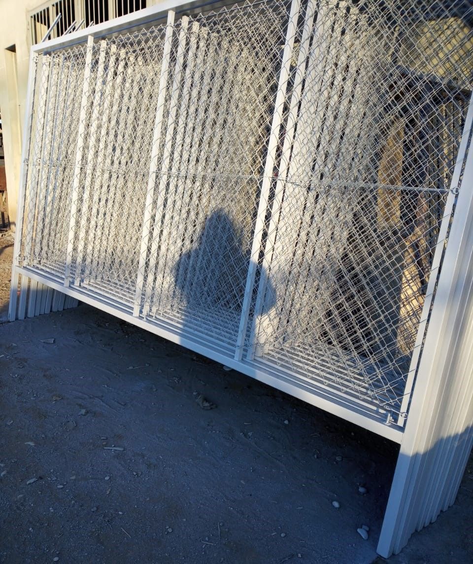 Valla metálica blanca con diseño de malla metálica, apoyada sobre hormigón. La sombra de una persona se proyecta sobre ella.