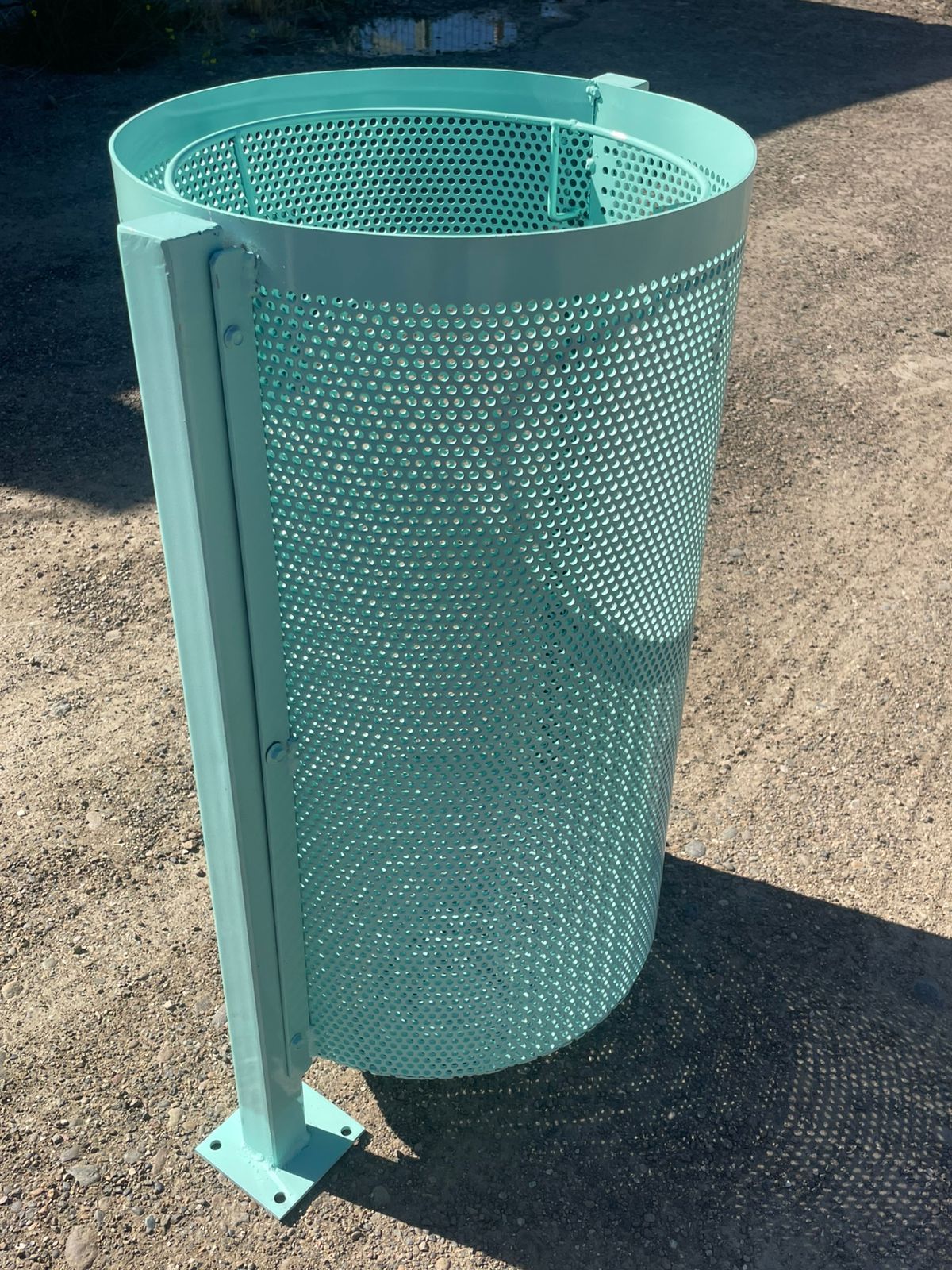 Un cubo de basura cilíndrico de metal turquesa con cuerpo perforado y poste cuadrado. Al aire libre, en un día soleado.