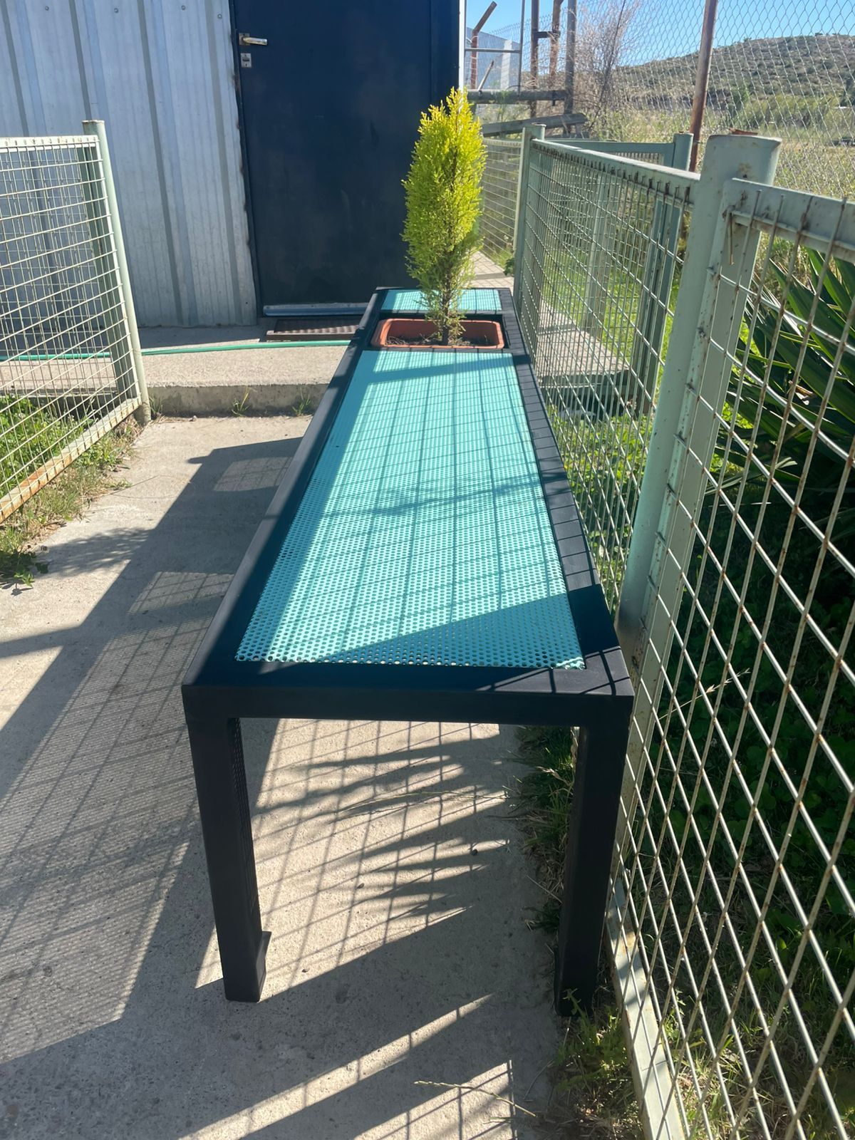 Mesa larga de color negro con tablero turquesa, un pequeño árbol y una valla metálica.