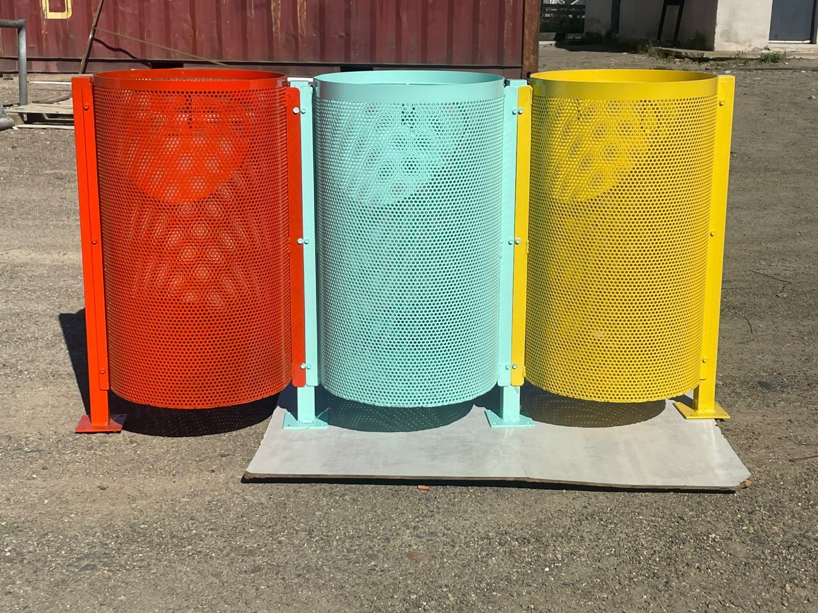 Tres contenedores de reciclaje de colores: rojo, verde azulado y amarillo, sobre una base de hormigón.