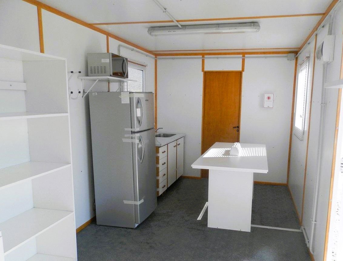 Cocina en un pequeño edificio portátil, con refrigerador, estantes, microondas y mesa pequeña.