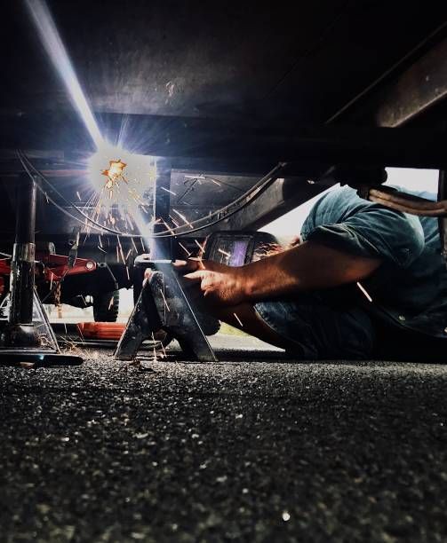 Person welding under a dark structure, wearing a mask, sparks flying, low angle shot.