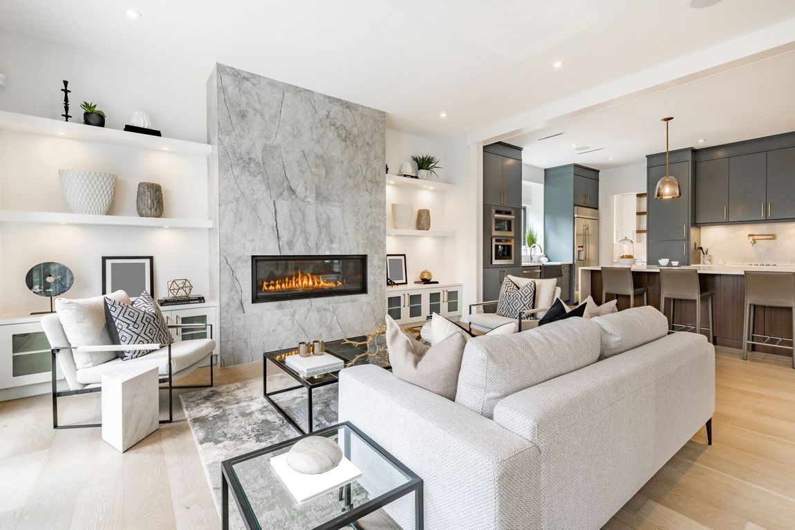 Bright modern living room with gray sectional, fireplace, white shelving, and open kitchen.