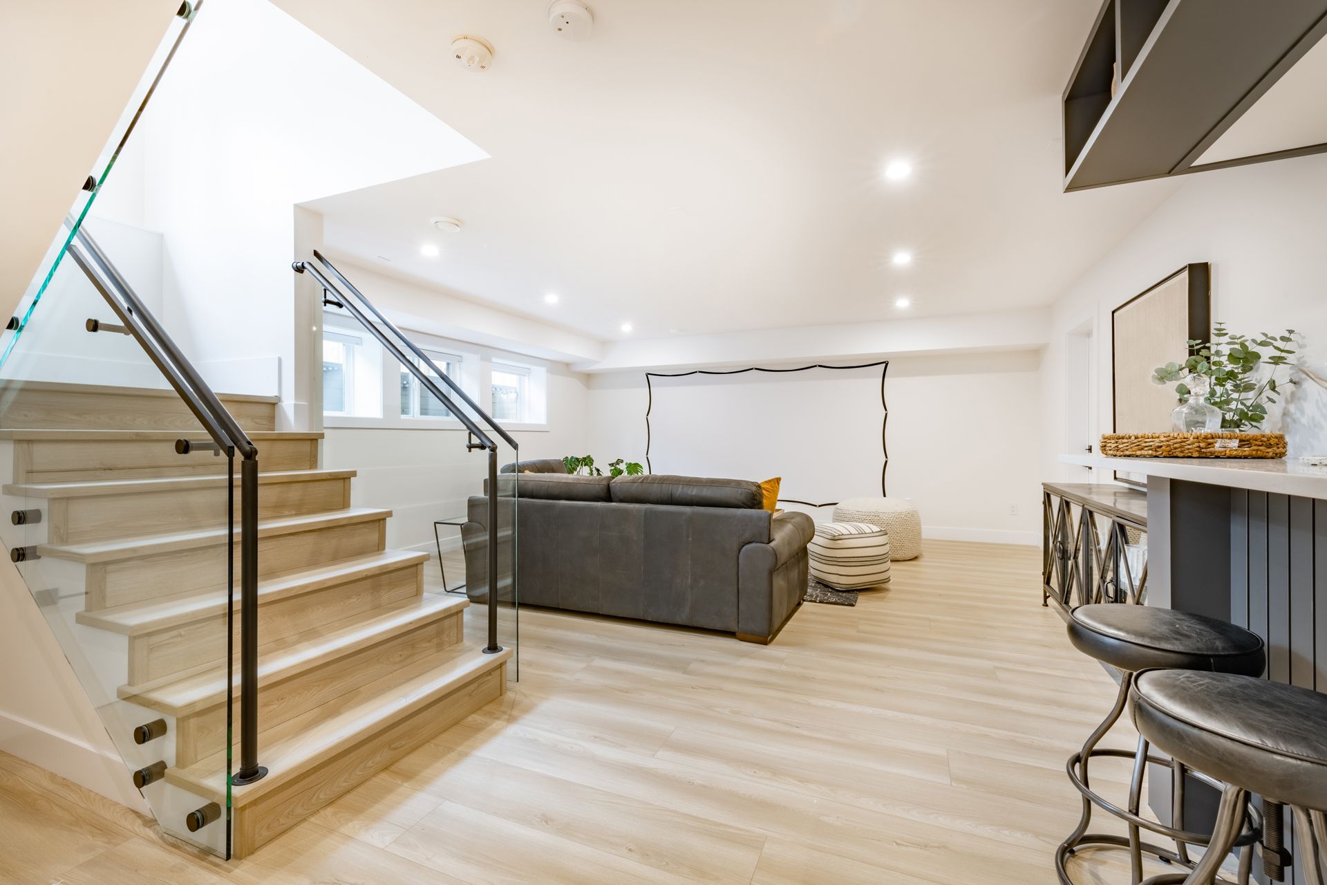 Modern open-plan living room with staircase, gray sofa, light wood floors, and a kitchen bar with stools