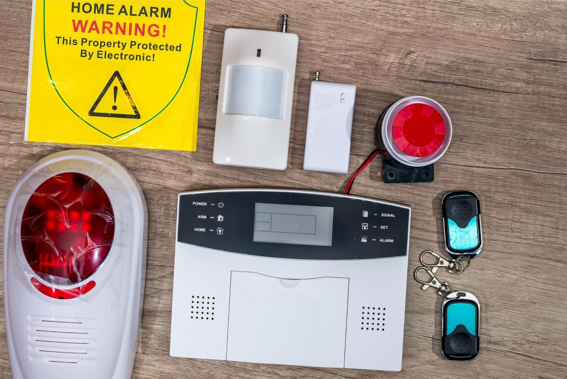 Home security alarm components on a table, including sensors, siren, remote controls, and warning label.