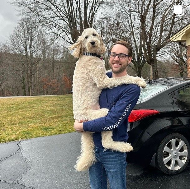 A man is holding a small dog in his arms in front of a car.