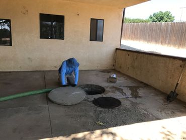 Worker in blue bends over a circular manhole cover in a concrete yard beside a building.