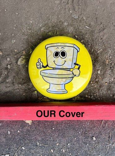 Yellow button with a cartoon toilet and the text “OUR Cover” on a pavement surface