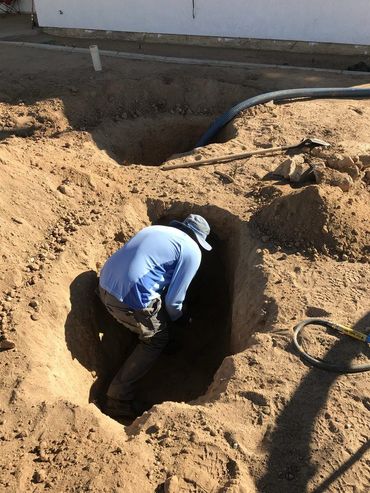 Worker in blue clothing crouches in a deep sandy trench at a construction site.