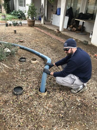 Man kneeling by a blue hose in a yard, working on a black drainage pipe near a house.