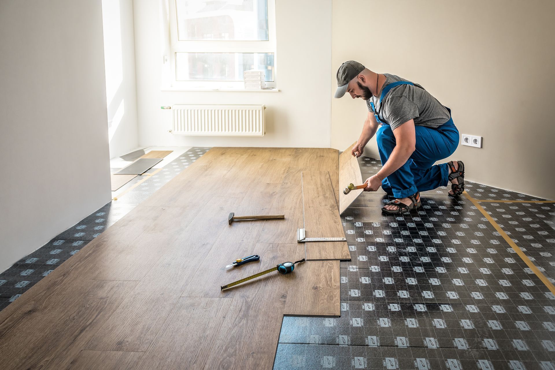 Person in blue overalls installing wood flooring in a room with tools laid out.