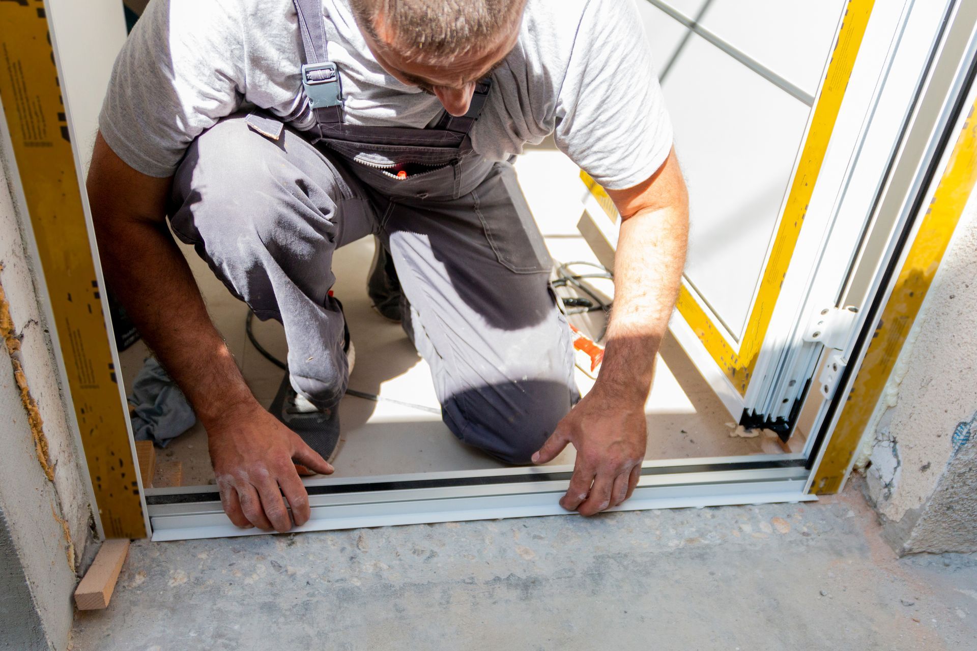 Man kneeling, installing a sliding door's bottom track in a doorway. White and yellow frame, concrete floor.