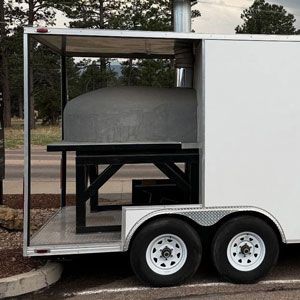 Pizza oven on a trailer, white exterior, black metal table, parked outdoors.