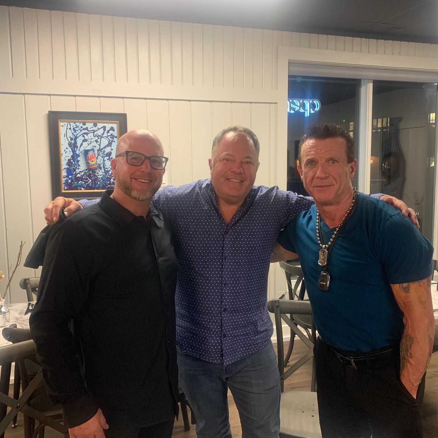 Three men indoors posing together; two have arms around the middle man. Restaurant setting.