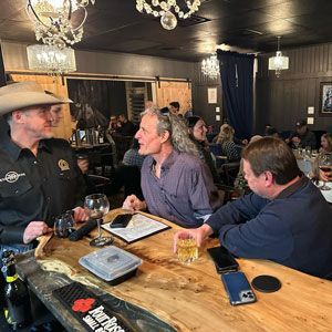 Three men at a bar. One in a cowboy hat, two others seated. Wooden bar, drinks, people in background.