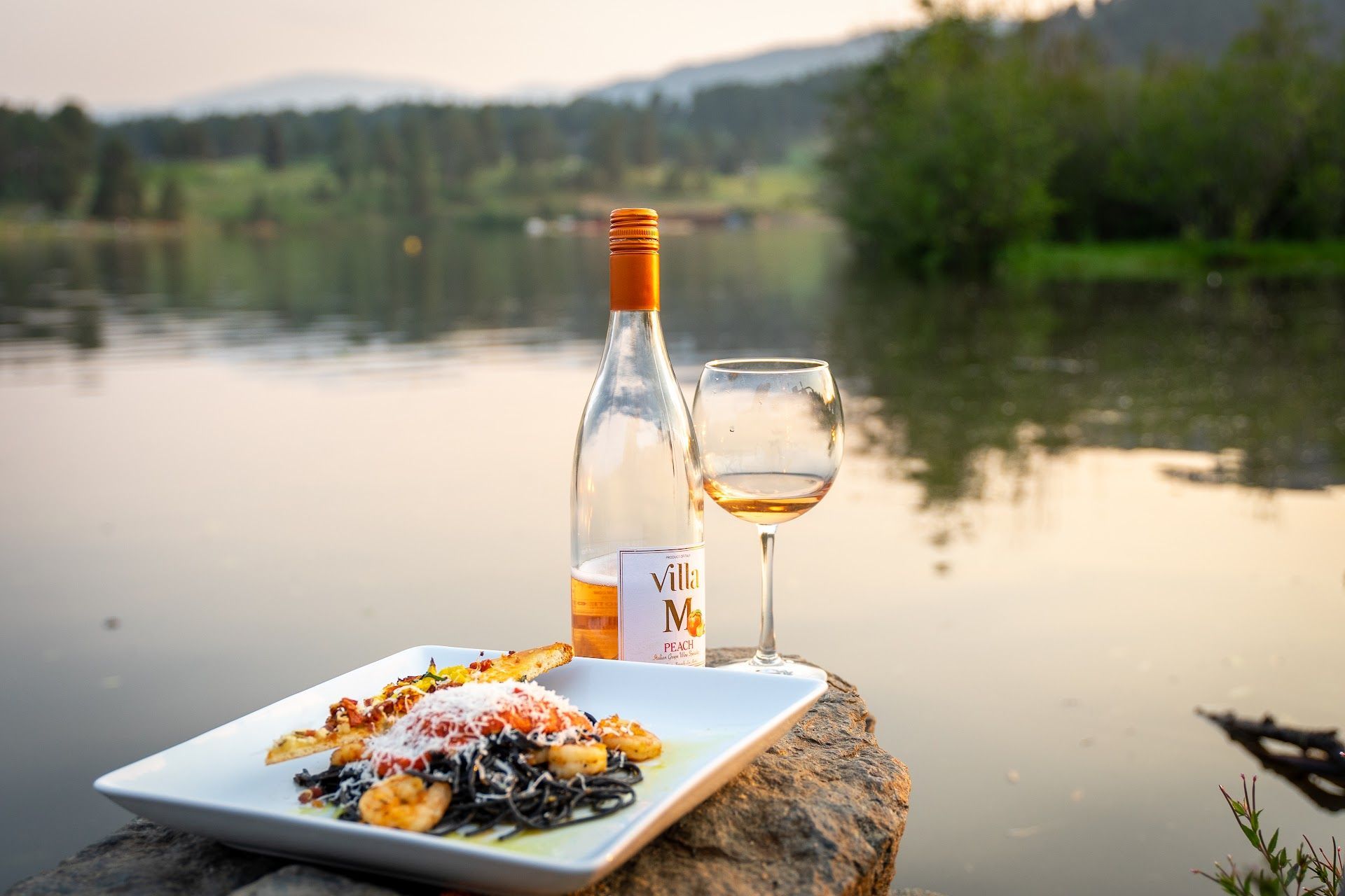 Bottle of wine, glass, and plate of pasta on a rock by a lake with forest background.