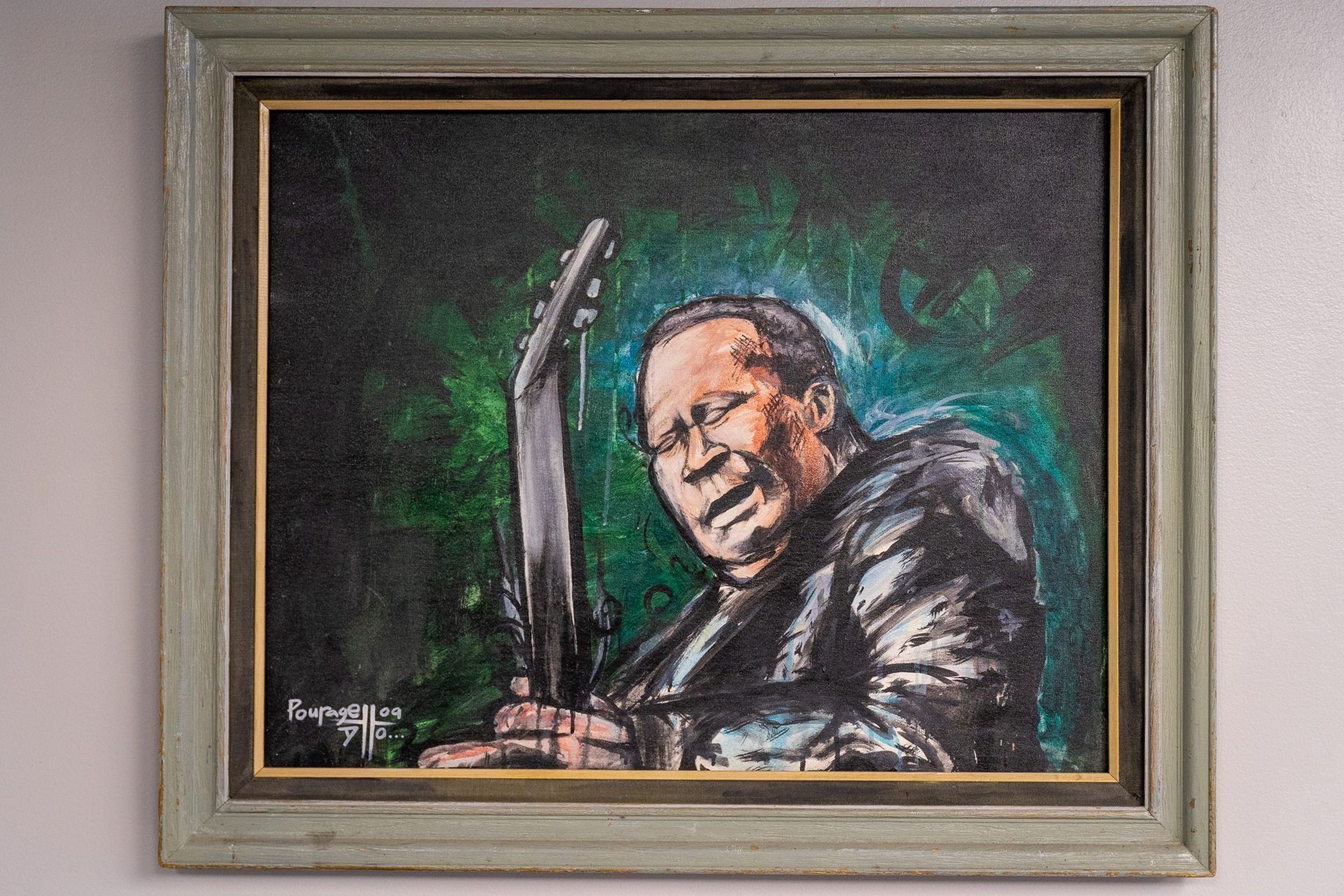 Painting of a musician holding a guitar, against a green and dark background; framed.