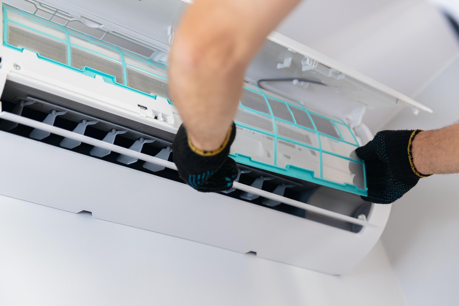 A technician wearing black gloves removes the air filter from a white wall-mounted air conditioner unit.