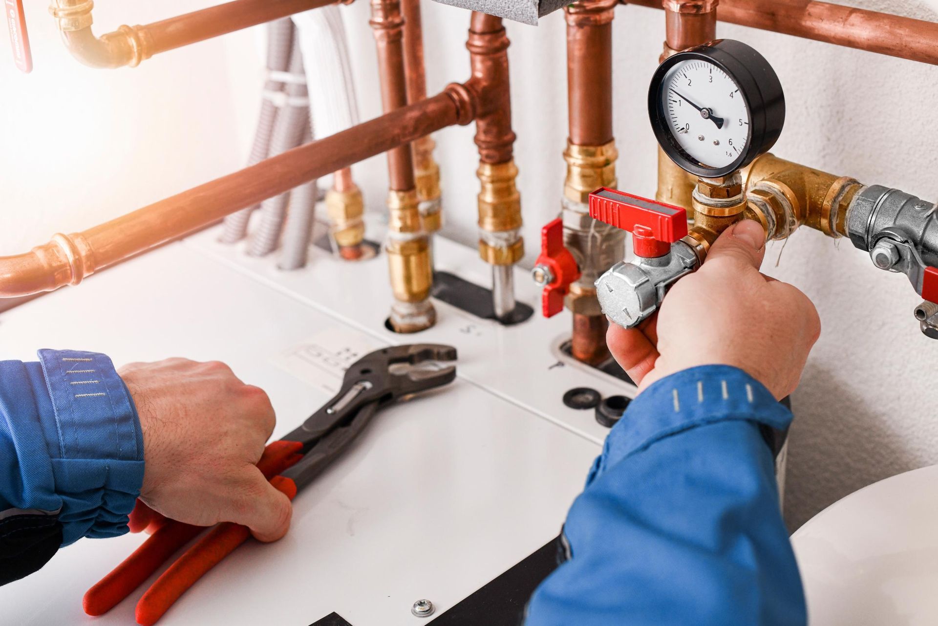 A person in blue workwear uses pliers to service copper pipes and a pressure gauge on a heating system.