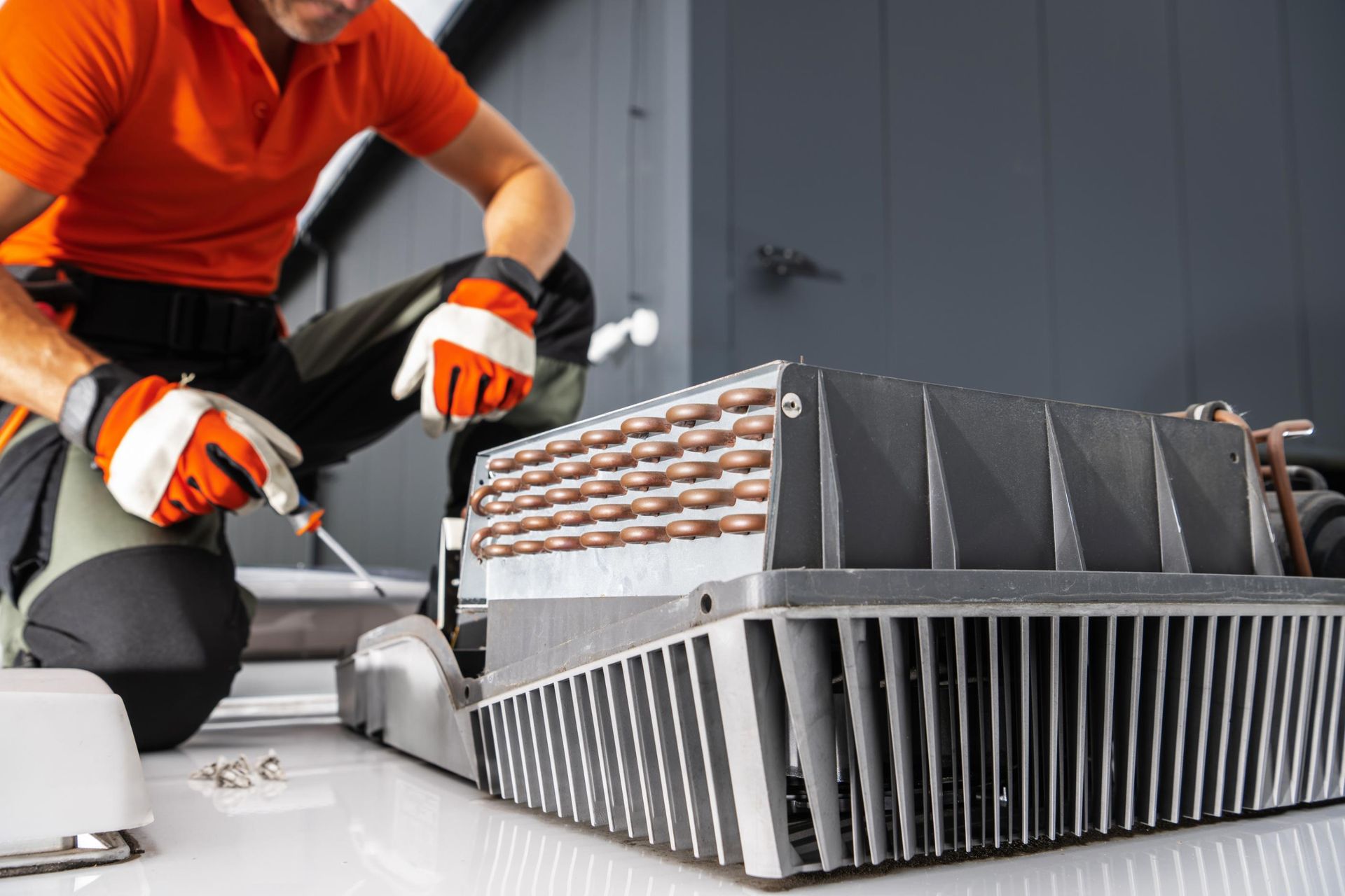 Technician in orange shirt and safety gloves using a screwdriver to repair a rooftop HVAC unit.