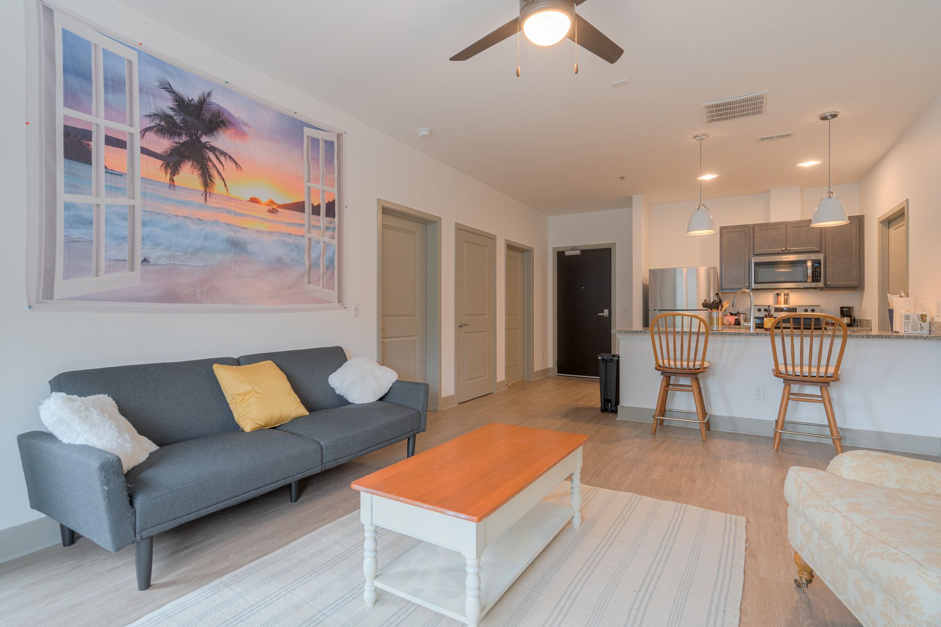 Living room with sofa, coffee table, and kitchen in the background. Beach scene artwork on the wall.