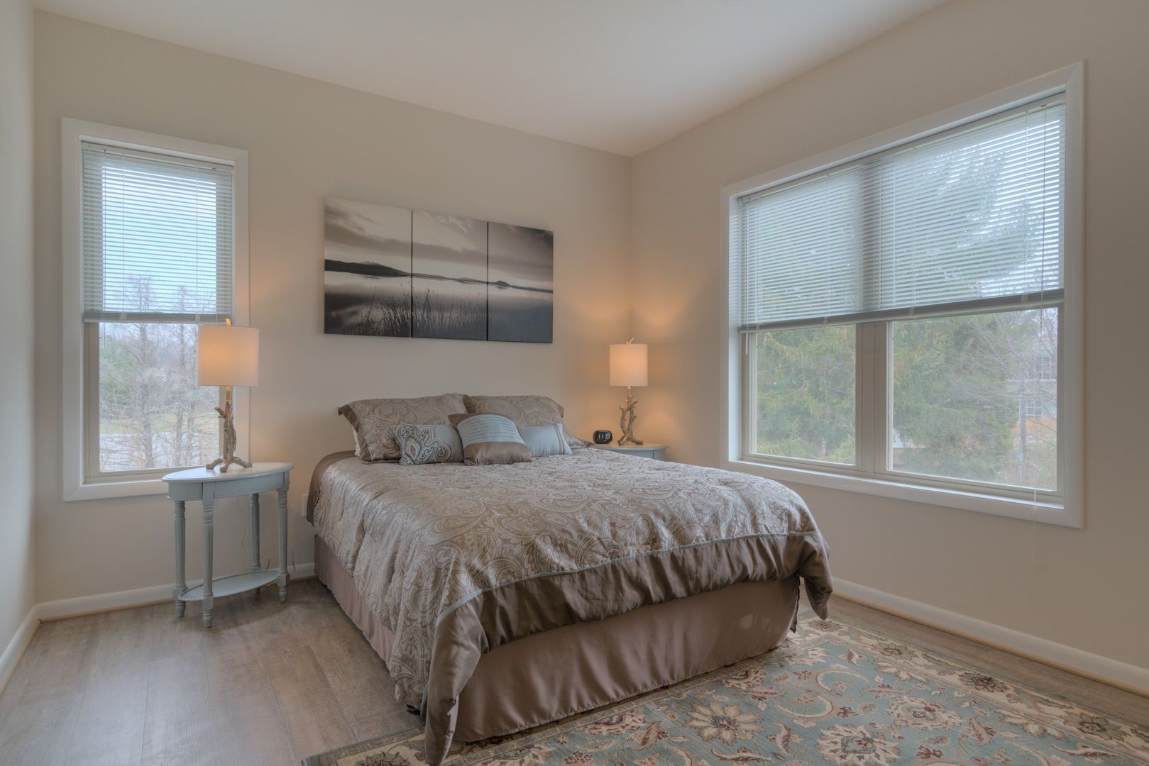 Bedroom with bed, windows with blinds, artwork, and side tables with lamps.