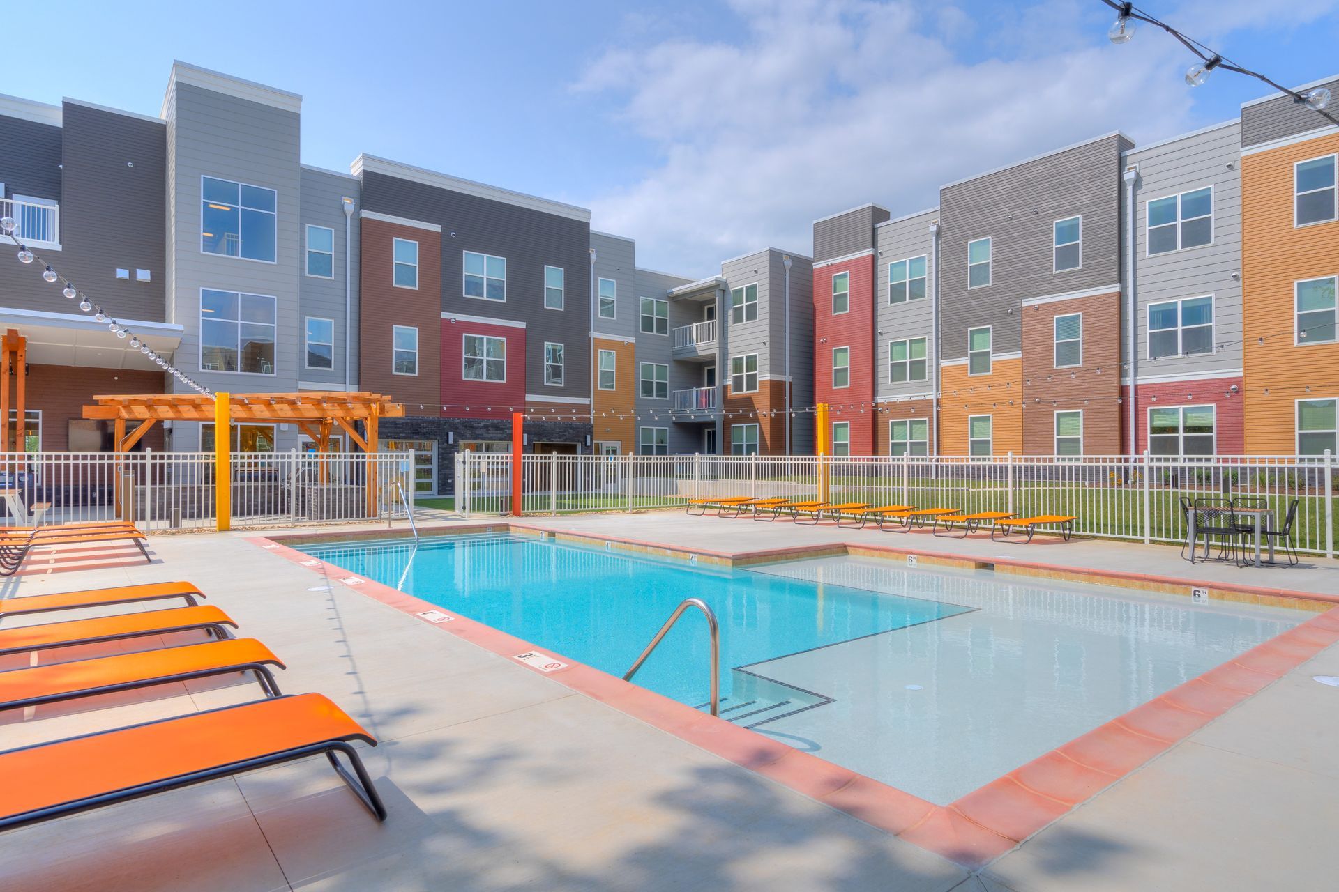 Pool area with orange lounge chairs, a fenced-in pool, and a multi-colored apartment building.