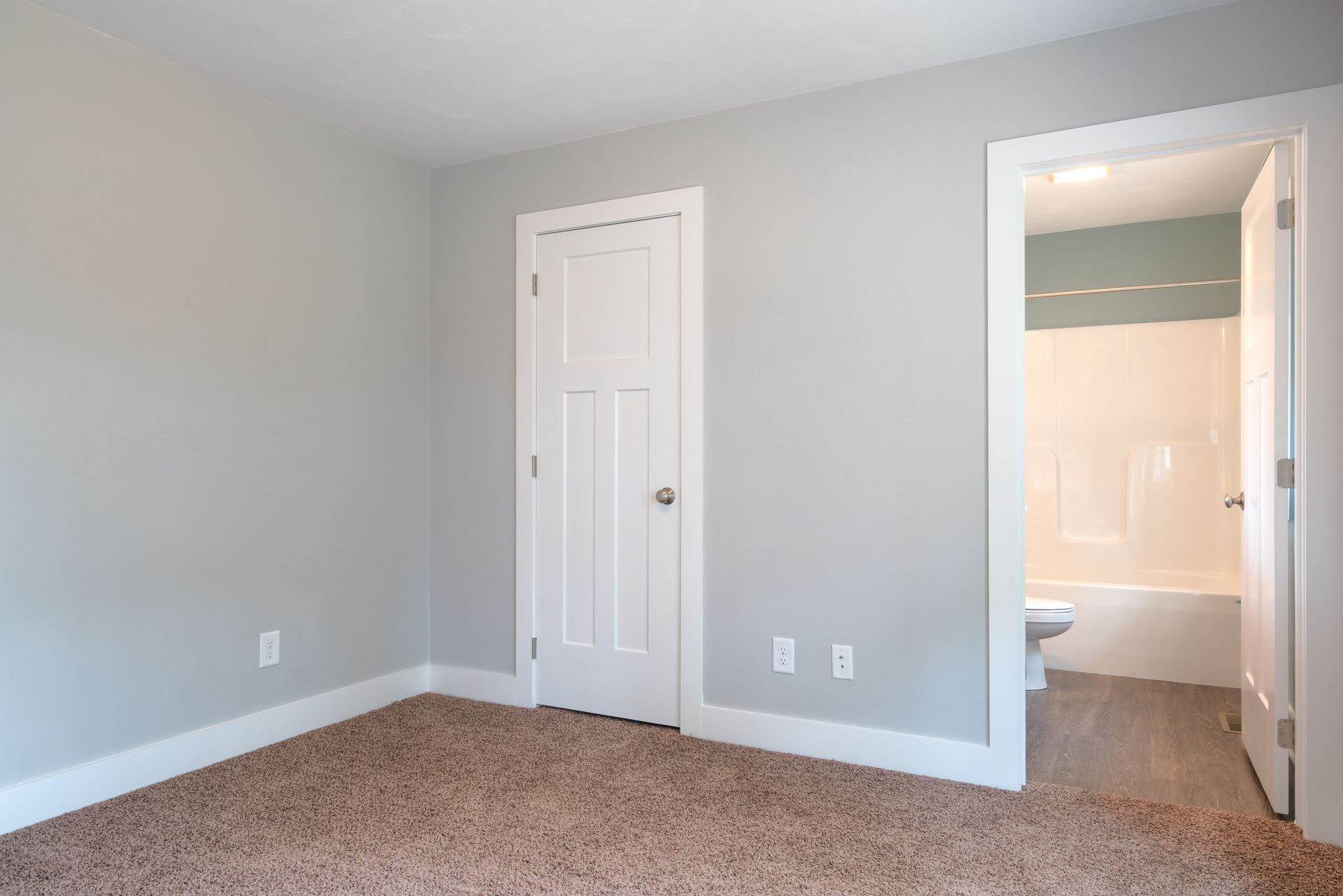 Empty bedroom with gray walls, white trim, and beige carpet. An open doorway leads to a bathroom.